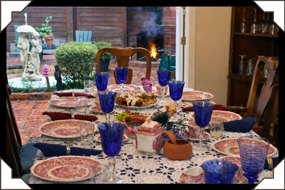 A beautifully set dining table with blue glassware and patterned plates, viewed through an open door to a garden.