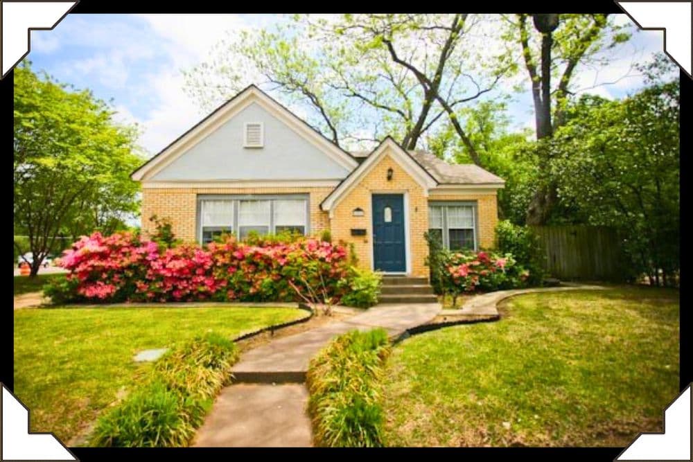 A charming yellow brick house surrounded by lush green trees and vibrant pink flower bushes.