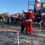 many people in background waiting to run along flagged pathway, in front of phot is "santa" and young man. Santa appears to be speaking to the crowd