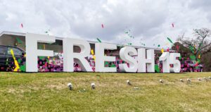 white letters in a grassy setting says Fresh15-has colorful flowers behind the letters