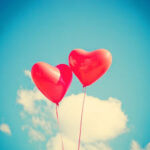 blue sky with fluffy white clouds. two red heart balloons in the foreground