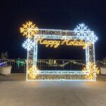 square frame of yellos and white lights overlooking the Tyler Rose garden there are 3 each of blue and yellow snowflake lights on the frame. The words Happy Hoidays is at the top in script