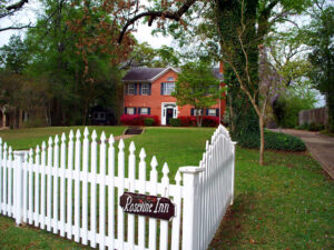 picture of Rosevine Inn red brick 2 story house in the background white picket fence in the forground with a sign on it that says Rosevine Inn long green lawn between the fence and the house